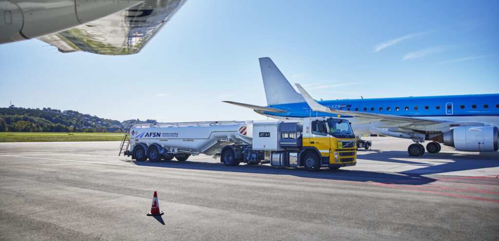 En flybane hvor man ser undersiden av en flyvinge og i midten av bildet ser man siden på en hvit og gul tankbil med AFSN logo på. Et hvitt og blått fly står bak tankbilen.